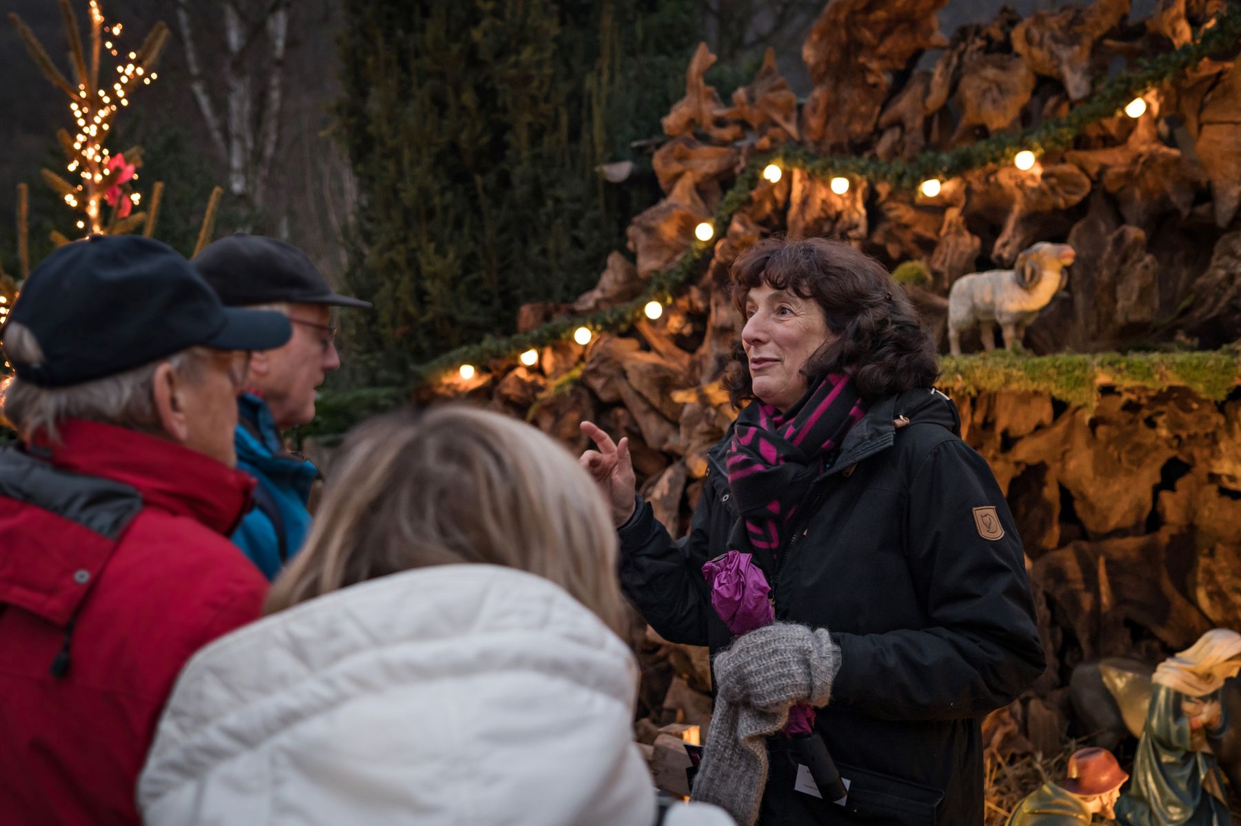 A woman speaks to three people in winter clothing near a festive outdoor display with lights, wooden decorations, and a sheep figure.